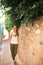 Caucasian girl in white T-shirt with fruits, green pants and gray sneakers is walking along a narrow ancient street with trees and