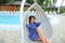 Caucasian happy woman sitting in pendant white chair, swiming pool in background.