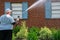 Caucasian heavy overweight man spraying water from a nozzle on the bricks and shutters on a house. There are bushes and plants in