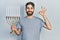Caucasian man with beard holding menorah hanukkah jewish candle doing ok sign with fingers, smiling friendly gesturing excellent