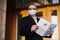 Caucasian man in sunglasses, medical mask and formal black suit, stands near building outdoor, holds newspaper in hands, poses at