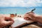 Caucasian man is writing sime idea, message or letter in his notepad by pen while he sitting on the beach of tropical