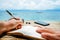 Caucasian man is writing sime idea, message or letter in his notepad by pen while he sitting on the beach of tropical
