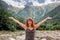 Caucasian middle aged woman holding arms up showing happiness being in valley between mountains