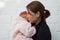 Caucasian middle aged woman holding on hands two years old child and looking aside with anger on the white brick background