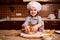 Caucasian mischievous little toddler girl rolling out dough on floured wooden board, smiling broadly looking at camera