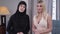 Caucasian and Muslim women looking at camera and smiling. Blond modern woman in candid dress and Muslim lady in black