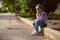 Caucasian pensive little kid girl in casual denim, sitting on paving stones, dreamily looking away, holding wild flower