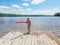 Caucasian preschool blonde girl in pink swimsuit standing on wooden lake river dock with pool noodle on summer day