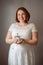 Caucasian red-haired pregnant woman in white dress standing at home holding small green plant in pot