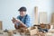 Caucasian senior old white bearded man carpenter in apron and hat working in workshop, holding and looking at handmade wood toy,