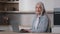 Caucasian senior teacher elderly housewife businesswoman grandmother grey-haired lady work on laptop in kitchen use home