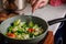 caucasian senior woman hand with fork picks frying vegetables in frying pan