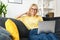 Caucasian senior woman spending home leisure with laptop. Calm woman sitting at cozy sofa and looking at camera. Web