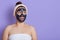 Caucasian surprised beautiful woman with towel on her body with black cleansing mask on face isolated over lilac background,