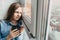 Caucasian teenage girl sits on windowsill, looks thoughtfully at the street outside the window and holds a black cup in her hands