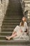 Caucasian white female model and brick stone. Beautiful girl, long red hair, beige skirt and cardigan. Woman siting on the stairs