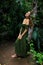 Caucasian woman leaning against a tree in tropical rain forest. Beautiful woman wearing long green dress. Walking trail in jungle