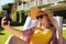 Caucasian woman sitting in sunny garden wearing sunhat and sunglasses taking selfie with smartphone