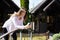 Caucasian woman surfing internet on modern smartphone while stand on garden bridge outdoors. Using mobile near wooden cabins