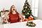 Caucasian young blonde woman sitting on the table by christmas tree crazy and mad shouting and yelling with aggressive expression