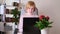 Caucasian young woman sits at laptop and types in cozy room. Nearby on table is houseplant.