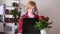 Caucasian young woman sits at laptop and types in cozy room. Nearby on table is houseplant.