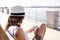 Caucasian young woman with white hat sitting on a bench of a sidewalk by the sea using her smartphone - Single woman outdoor