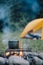 Cauldron over burning campfire with yellow tent on background green forest at warm evening. Bonfire cooking in summer or autumn