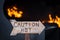 Caution warning hot wooden arrow sign with orange fire and flames burning in the background.