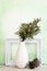 Cedar branch in a white vase against a painterly green background with empty picture frame and pine cones