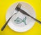 The celebration of April fool`s Day, a Plate with a fork and knife and a paper fish on a yellow colored background. Humor