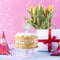 Celebratory cake with white cream for birthday and flowers, on a pink and blue background. Tinsel, waverki and boxes of gifts.