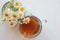 Chamomile herbal tea in glass cup and bouquet of white daisies in a glass vase on the white wooden background with copy space. Top