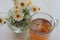 Chamomile herbal tea in glass cup and bouquet of white daisies in a glass vase on the white wooden background with copy space. Top