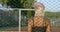 Charismatic blonde female is standing behind metal grid outdoors in sunny day, back view