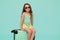 Charming baby girl in summer wear and trendy glasses, sitting on her turquoise plastic valise, isolated blue background