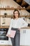 Charming brown-haired woman with long hair in white shirt and jeans stands against background of fragment of kitchen