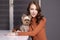 Charming brunette woman in brown golf with a Yorkshire Terrier puppy sitting at table in gray room. pet, animal