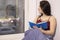 Charming brunette woman wrapped in blue blanket sitting on windowsill with blue notebook. morning routine, coziness, reading, city