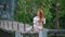 Charming cute young woman with huge shock of fiery red hair sits on suspension bridge over river and and braids her hair