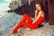 Charming elegant young woman in coral red one shoulder jumpsuit sitting on the beach at the old rusty piles