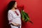 Charming ethnic pregnant woman in embroidered dress smelling bunch of tulips, gently caressing her belly, red background