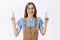 Charming friendly-looking charismatic female in glasses and trendy brown overalls pointing up with raised arms and