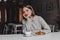 Charming girl with dark blond hair with smile looks into camera, posing at table in cafe with croissant and cup of