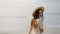 Charming girl in flowery dress posing, smiling at seaside, foamy water waves. Young brunette woman in straw hat walking