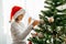 Charming girl in a santa claus hat decorates the Christmas tree at home with a garland with lights. Preparing for the