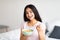 Charming Indian lady eating tasty cereal with milk, enjoying nutritious cornflakes in bedroom