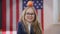 Charming joyful teen schoolgirl looking at camera smiling with apple on head and USA flag at background. Portrait of