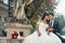 Charming loving just married couple is hugging and sitting on the fountains near the wedding bouquet of red flowers.